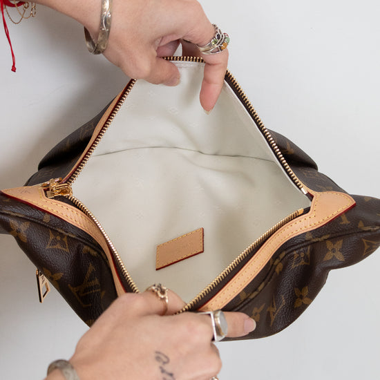 A Louis Vuitton monogram shoulder bag in brown coated canvas with tan leather trim, shown open with hands holding the zipper area. The interior is lined with light beige fabric, and a small tan leather brand patch is visible inside.
