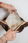 A Louis Vuitton monogram shoulder bag in brown coated canvas with tan leather trim, shown open with hands holding the zipper area. The interior is lined with light beige fabric, and a small tan leather brand patch is visible inside.