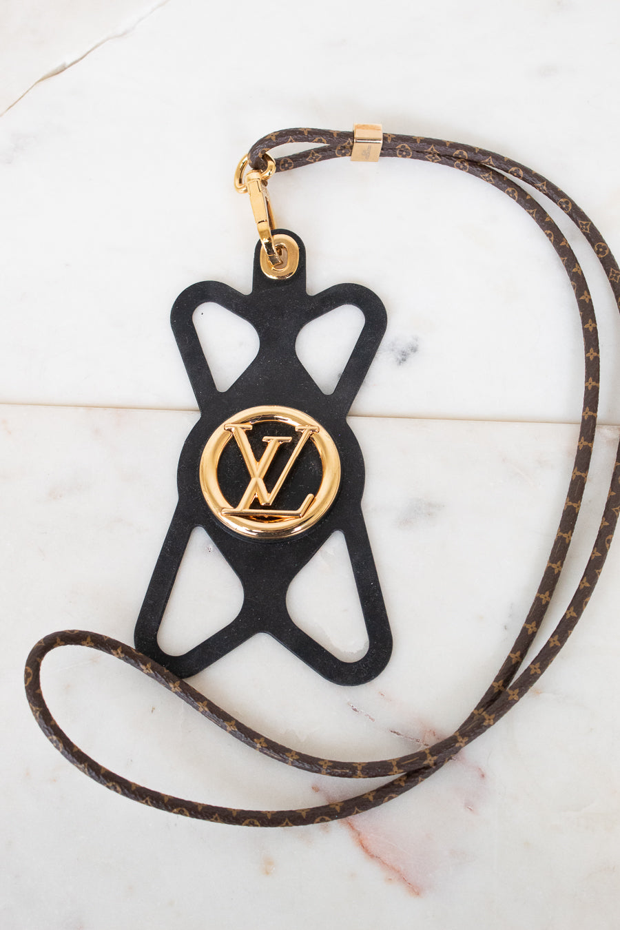 A Louis Vuitton monogram leather strap with a black silhouette-shaped pendant featuring a gold-tone Louis Vuitton LV logo medallion, displayed on a light marble surface.