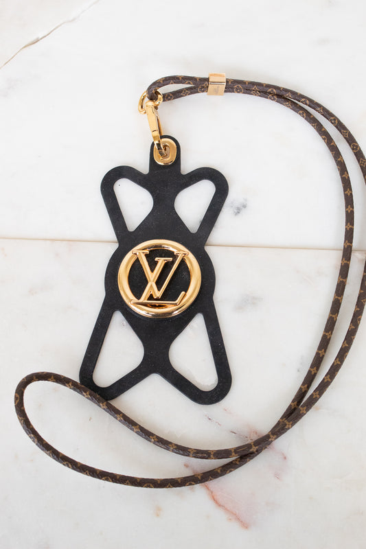 A Louis Vuitton monogram leather strap with a black silhouette-shaped pendant featuring a gold-tone Louis Vuitton LV logo medallion, displayed on a light marble surface.