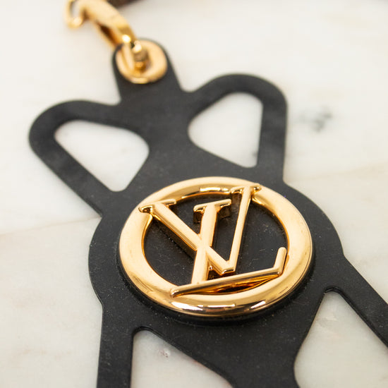 A Louis Vuitton keychain featuring a black leather silhouette of the brand's monogram with a polished gold-tone metal LV logo in the center, attached to a brown and gold patterned leather strap.
