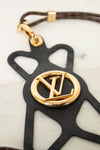 A Louis Vuitton keychain featuring a black leather silhouette of the brand's monogram with a polished gold-tone metal LV logo in the center, attached to a brown and gold patterned leather strap.