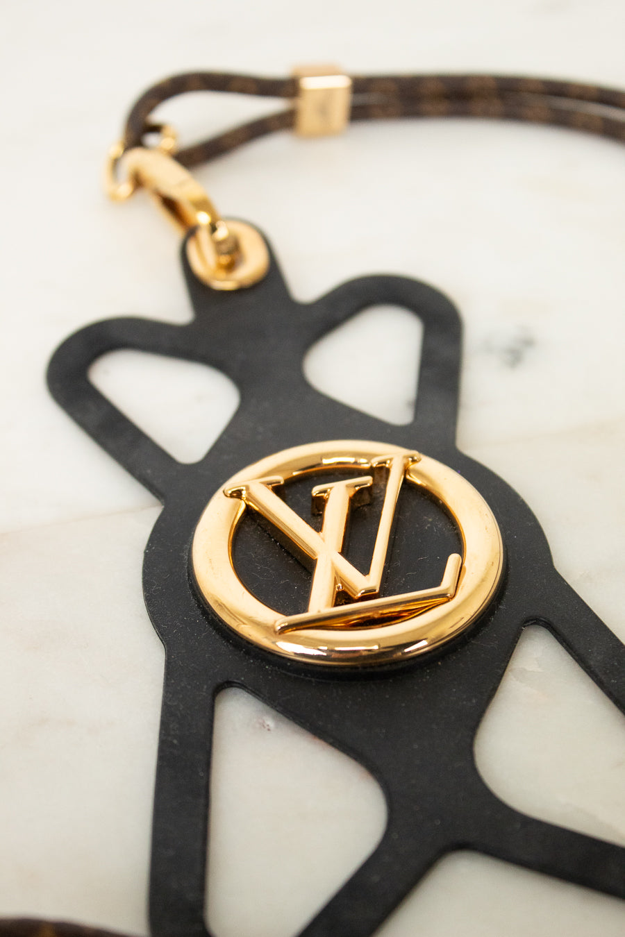 A Louis Vuitton keychain featuring a black leather silhouette of the brand's monogram with a polished gold-tone metal LV logo in the center, attached to a brown and gold patterned leather strap.