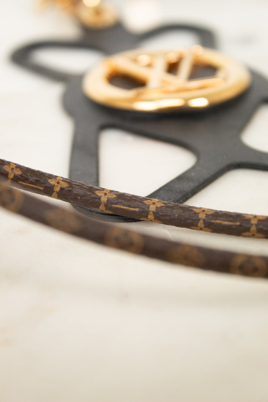 Close-up of a Louis Vuitton monogram canvas strap with gold-tone hardware and a black monogram-shaped plaque on a light marble surface.