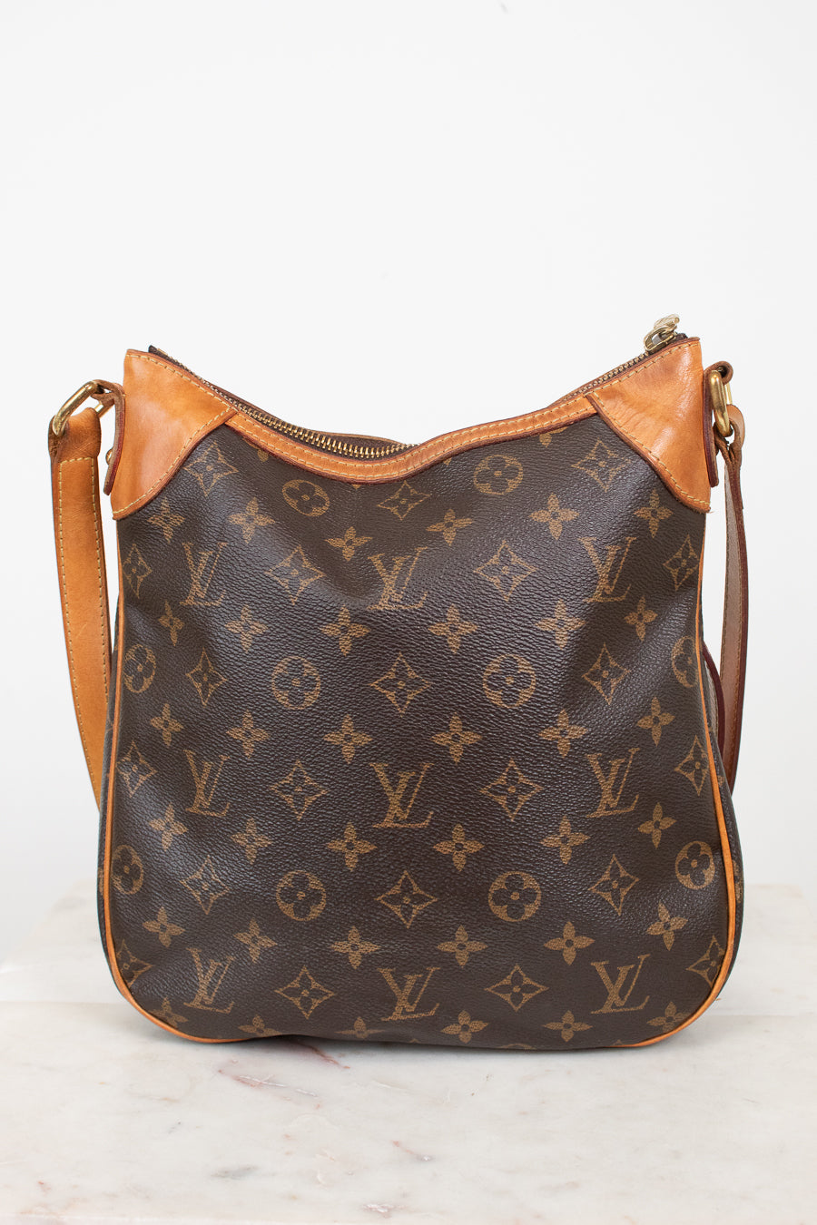 A Louis Vuitton monogram canvas shoulder bag with tan leather trim and a gold-tone zipper, displayed on a light marble surface against a white background.