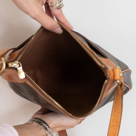 A close-up of a person holding open a brown monogram shoulder bag with tan leather trim and gold-tone zipper hardware, showing the interior against a plain light background.