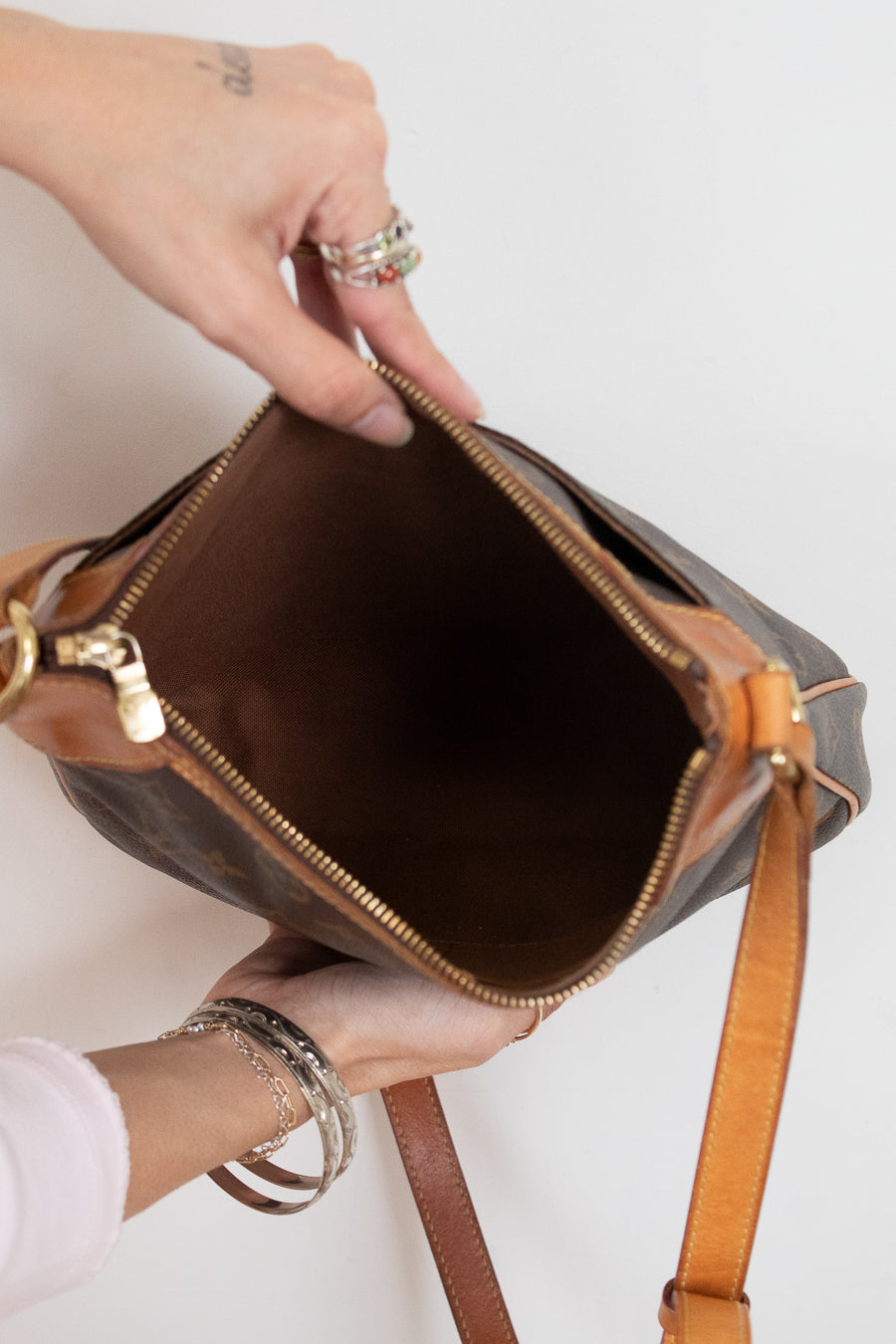 A close-up of a person holding open a brown monogram shoulder bag with tan leather trim and gold-tone zipper hardware, showing the interior against a plain light background.