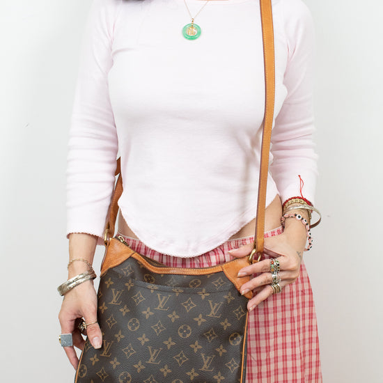 A woman wearing a white long-sleeve top and a red-and-white gingham skirt carries a brown Louis Vuitton monogram shoulder bag with tan leather trim and strap against a plain white wall.