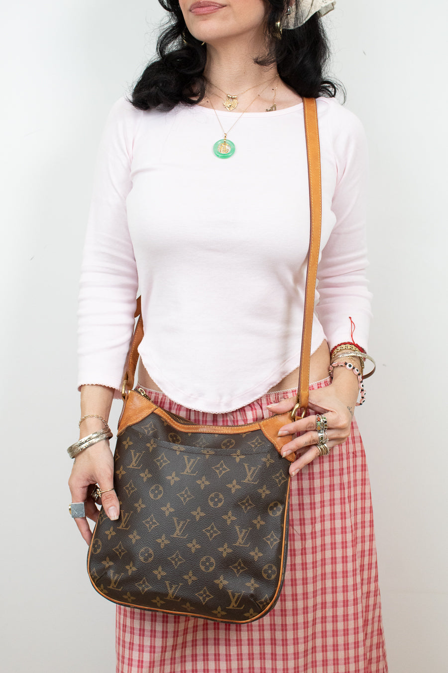A woman wearing a white long-sleeve top and a red-and-white gingham skirt carries a brown Louis Vuitton monogram shoulder bag with tan leather trim and strap against a plain white wall.