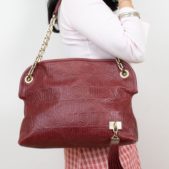 A woman wearing a white long-sleeve top and a red-and-white gingham skirt carries a large Louis Vuitton shoulder bag in burgundy red textured leather with gold-tone chain and tassel details.