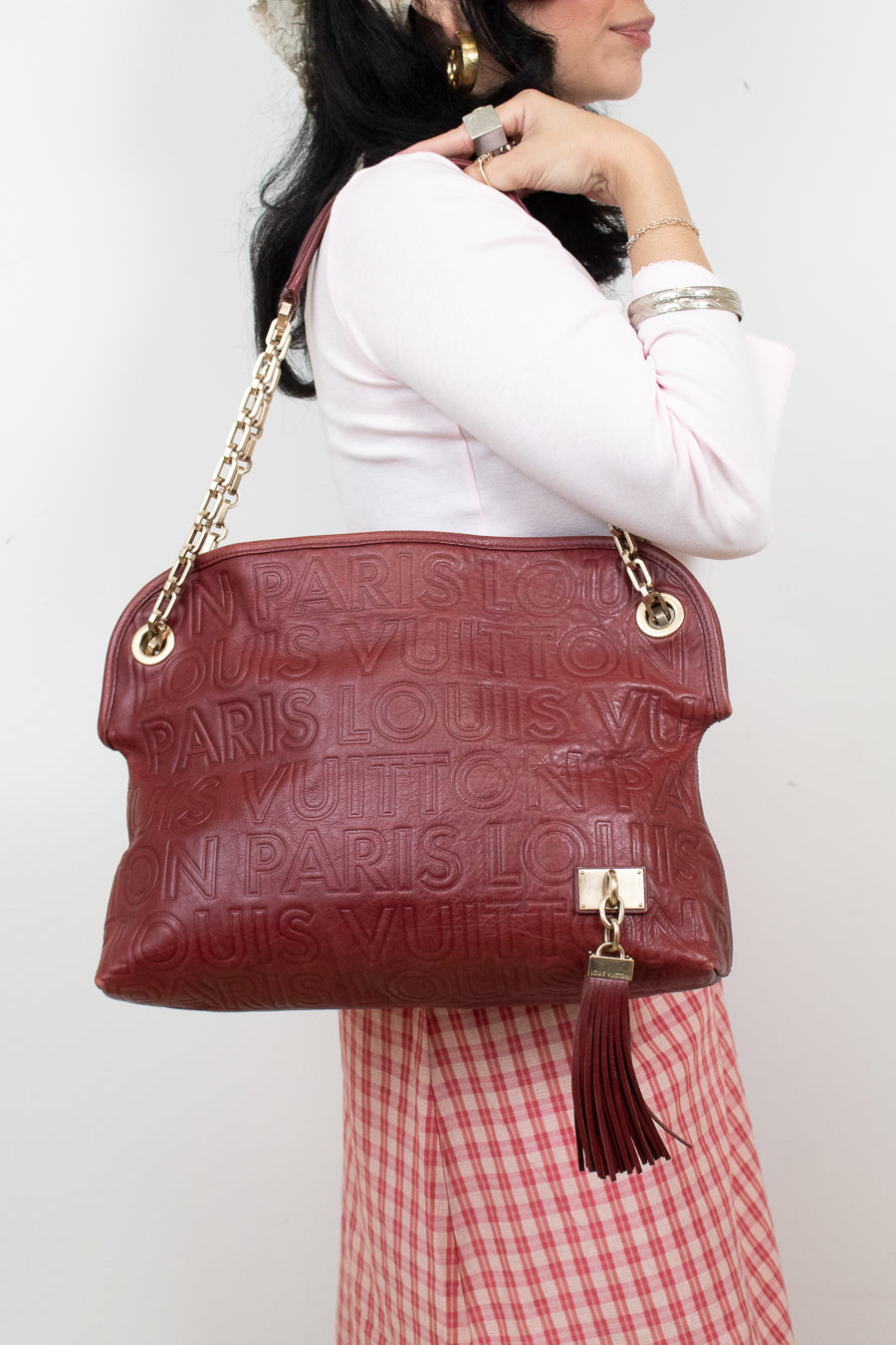 A woman wearing a white long-sleeve top and a red-and-white gingham skirt carries a large Louis Vuitton shoulder bag in burgundy red textured leather with gold-tone chain and tassel details.