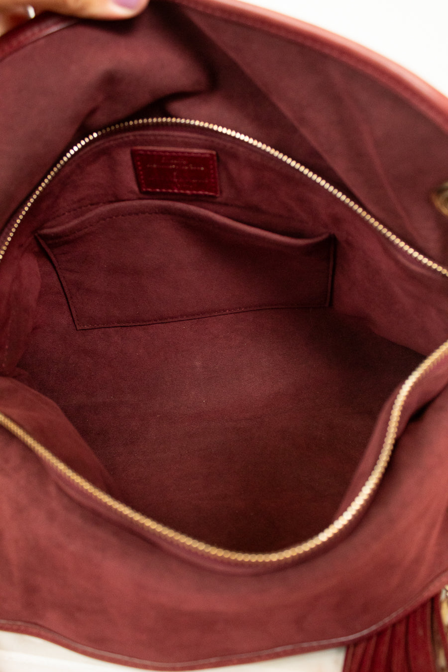 Close-up of the interior of a burgundy suede handbag, showing a zippered interior with a small pocket and a small leather brand patch inside.