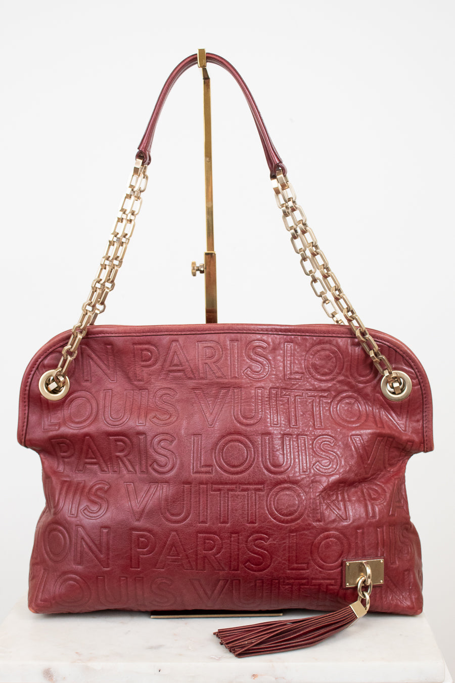 A Louis Vuitton monogram embossed leather shoulder bag in burgundy red, featuring gold-tone chain handles and a tassel detail, displayed on a white background.