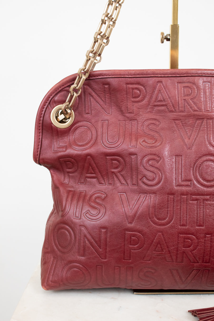 A close-up of a Louis Vuitton shoulder bag in burgundy leather with embossed 'LOUIS VUITTON PARIS' text, featuring a gold-tone chain strap and a gold metal ring attachment.