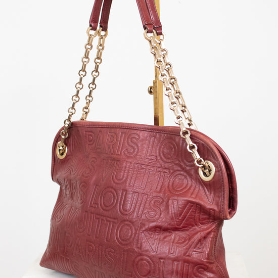A Louis Vuitton shoulder bag in embossed burgundy leather, featuring gold-tone chain straps, a top handle, and a tassel detail, displayed on a white marble surface against a white background.