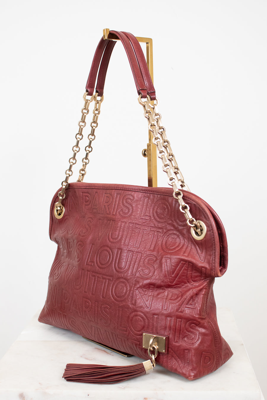 A Louis Vuitton shoulder bag in embossed burgundy leather, featuring gold-tone chain straps, a top handle, and a tassel detail, displayed on a white marble surface against a white background.