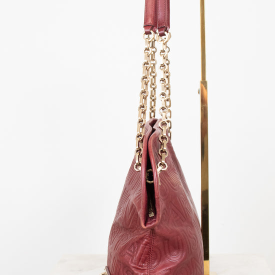 A burgundy red leather shoulder bag with embossed logo detailing, gold-tone chain straps, and a matching leather tassel, displayed on a gold stand against a white background.