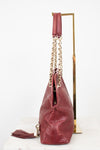 A burgundy red leather shoulder bag with embossed logo detailing, gold-tone chain straps, and a matching leather tassel, displayed on a gold stand against a white background.