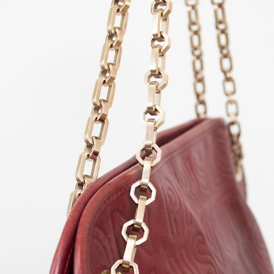 Close-up of a burgundy embossed leather handbag with a gold-tone chain strap against a white background.