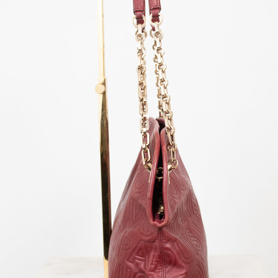 A burgundy leather shoulder bag with embossed logo detailing and gold-tone chain straps, displayed on a gold stand against a white background.