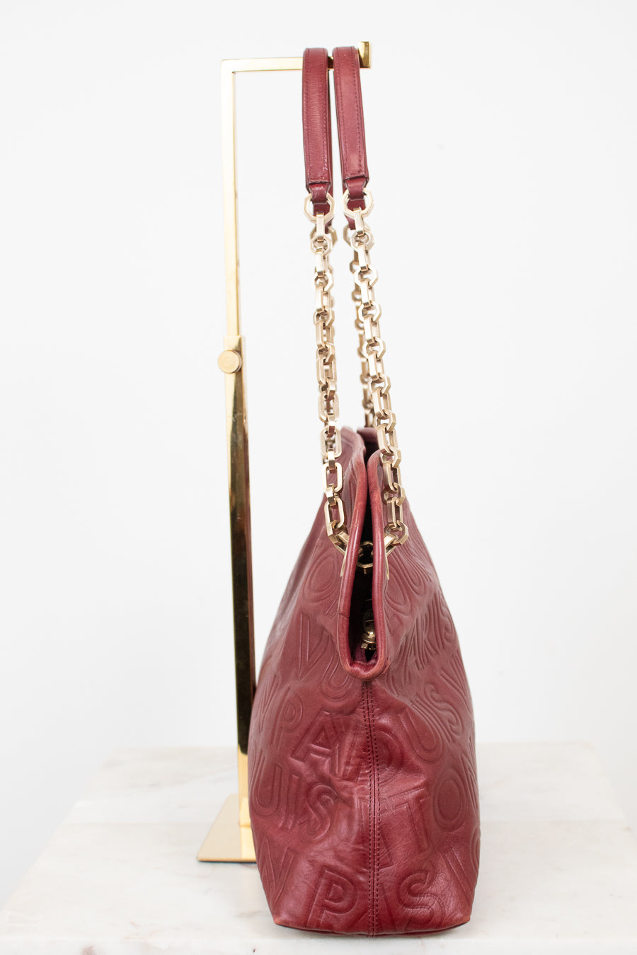 A burgundy leather shoulder bag with embossed logo detailing and gold-tone chain straps, displayed on a gold stand against a white background.