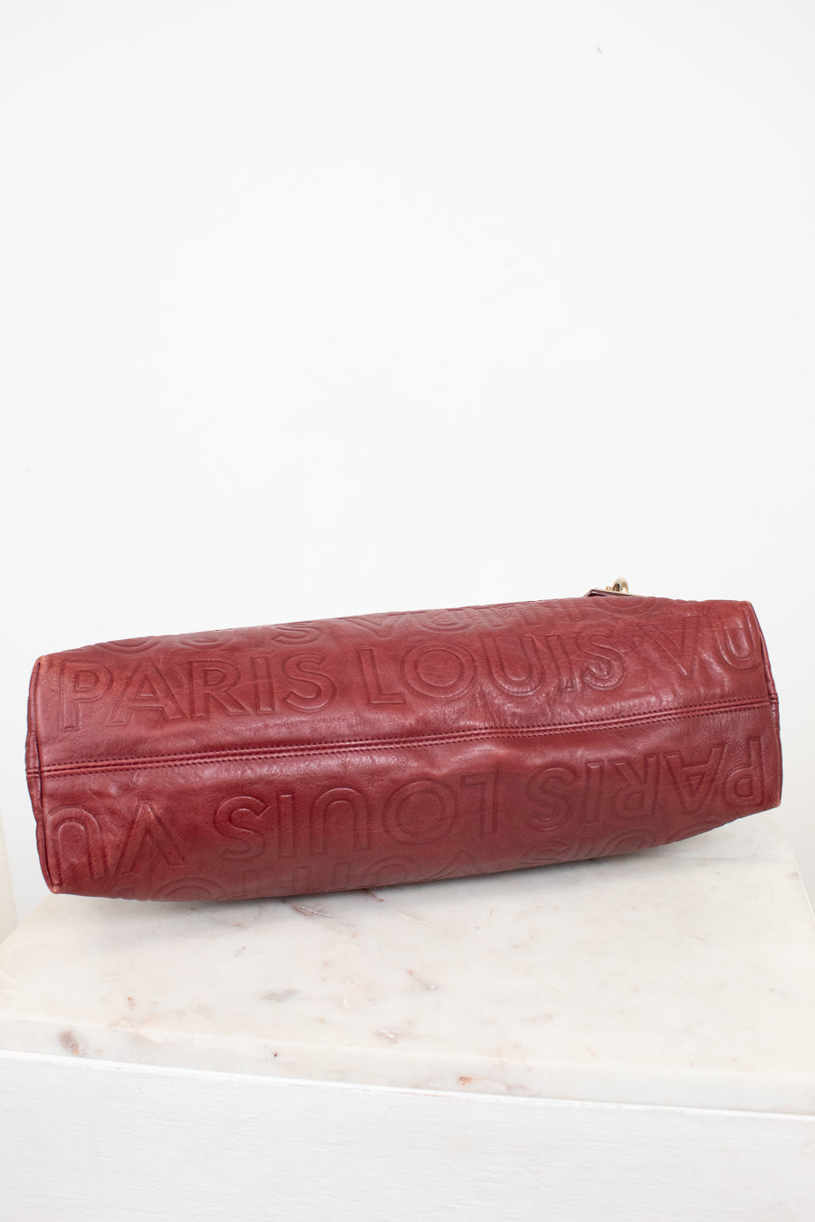 A Louis Vuitton burgundy leather shoulder bag with an embossed monogram pattern, resting on a white marble surface against a white background.