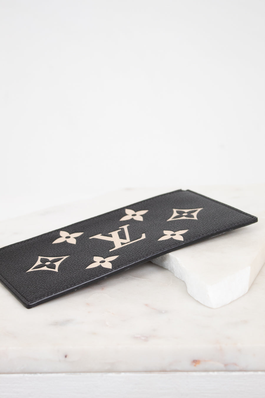 A black leather Louis Vuitton monogram patch with gold-tone LV logos and floral motifs, resting on a white marble block against a white background.
