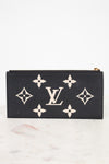 A black Louis Vuitton zip-around wallet or pouch made of textured leather, featuring the classic cream monogram pattern and LV logo on the front, set against a white background on a light marble surface.