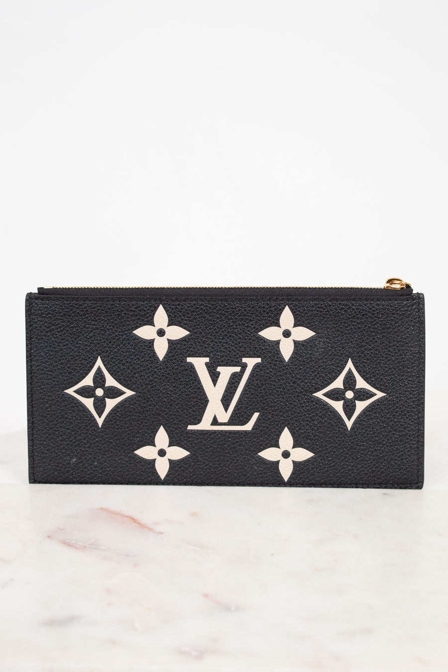 A black Louis Vuitton zip-around wallet or pouch made of textured leather, featuring the classic cream monogram pattern and LV logo on the front, set against a white background on a light marble surface.