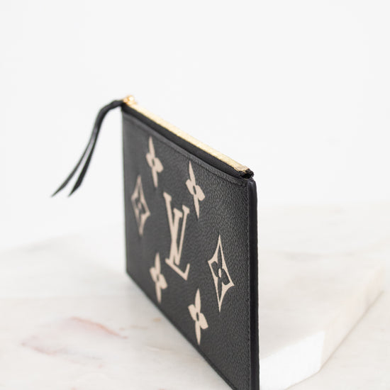 A black textured leather Louis Vuitton pouch featuring the classic beige monogram pattern, shown standing on a white marble surface against a white background.