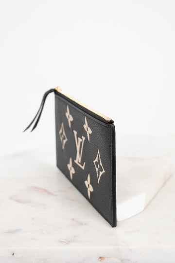 A black textured leather Louis Vuitton pouch featuring the classic beige monogram pattern, shown standing on a white marble surface against a white background.