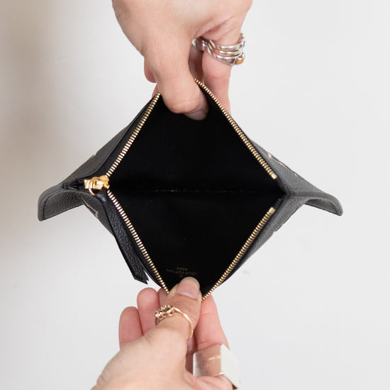 A close-up of a black leather zip-around card holder or small wallet being opened by two hands, showing a black interior and gold-tone zipper hardware against a plain light background.