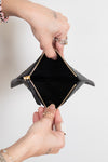 A close-up of a black leather zip-around card holder or small wallet being opened by two hands, showing a black interior and gold-tone zipper hardware against a plain light background.