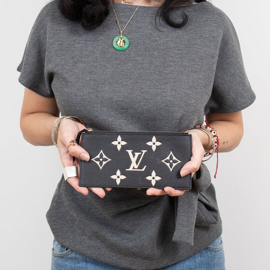 A woman wearing a gray t-shirt and blue jeans holds a black Louis Vuitton monogram pouch against a plain light gray background.