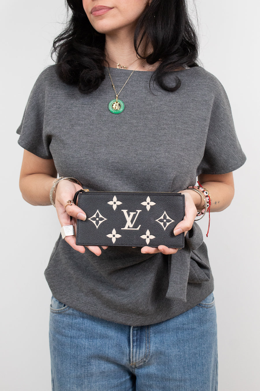 A woman wearing a gray t-shirt and blue jeans holds a black Louis Vuitton monogram pouch against a plain light gray background.