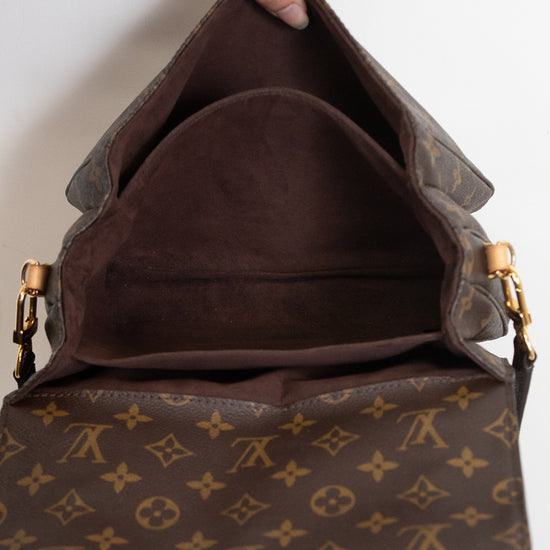 A Louis Vuitton monogram shoulder bag shown open, with a hand holding the top flap to display the interior. The bag features the classic brown and tan monogram canvas with gold-tone hardware.