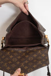 A Louis Vuitton monogram shoulder bag shown open, with a hand holding the top flap to display the interior. The bag features the classic brown and tan monogram canvas with gold-tone hardware.
