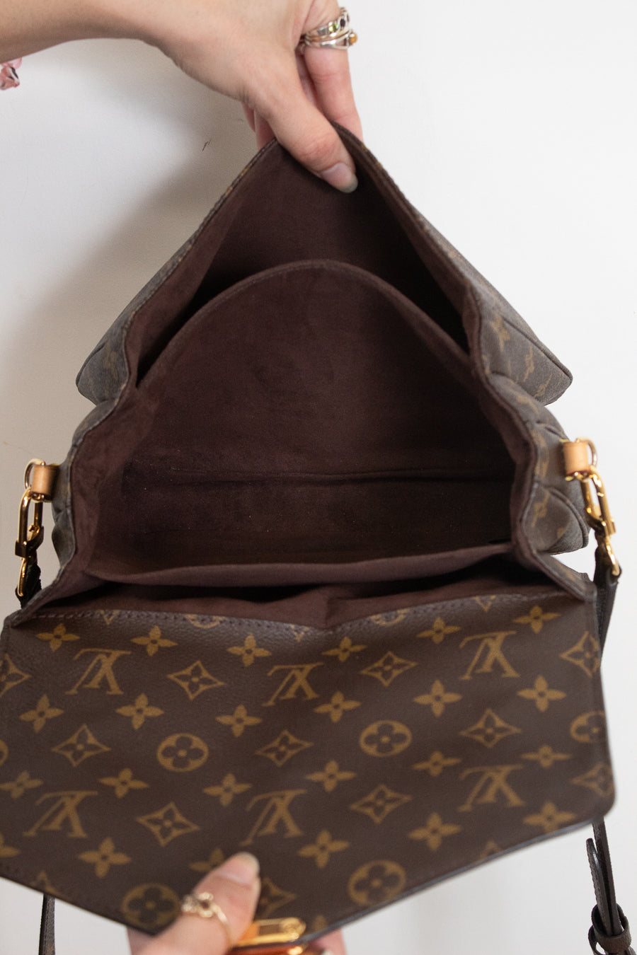 A Louis Vuitton monogram shoulder bag shown open, with a hand holding the top flap to display the interior. The bag features the classic brown and tan monogram canvas with gold-tone hardware.