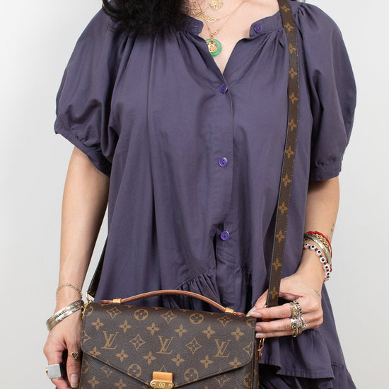 A woman wearing a purple puff-sleeve button-up dress holds a Louis Vuitton monogram shoulder bag with a brown leather handle and strap against a plain light background.