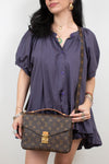 A woman wearing a purple puff-sleeve button-up dress holds a Louis Vuitton monogram shoulder bag with a brown leather handle and strap against a plain light background.