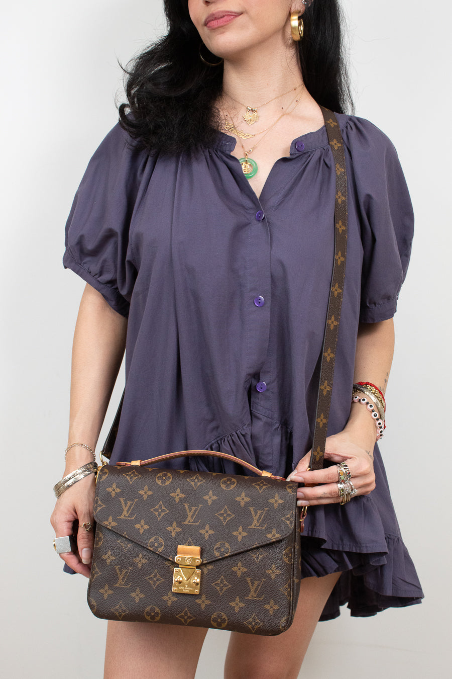 A woman wearing a purple puff-sleeve button-up dress holds a Louis Vuitton monogram shoulder bag with a brown leather handle and strap against a plain light background.