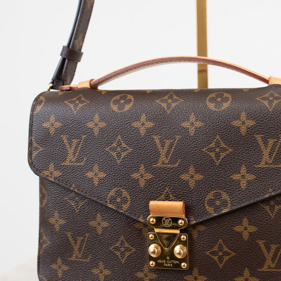 Louis Vuitton Monogram handbag in brown coated canvas with gold-tone hardware, displayed on a marble surface with a brass stand against a white background.