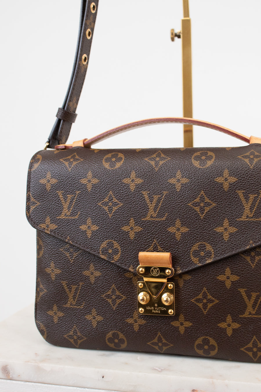 Louis Vuitton Monogram handbag in brown coated canvas with gold-tone hardware, displayed on a marble surface with a brass stand against a white background.