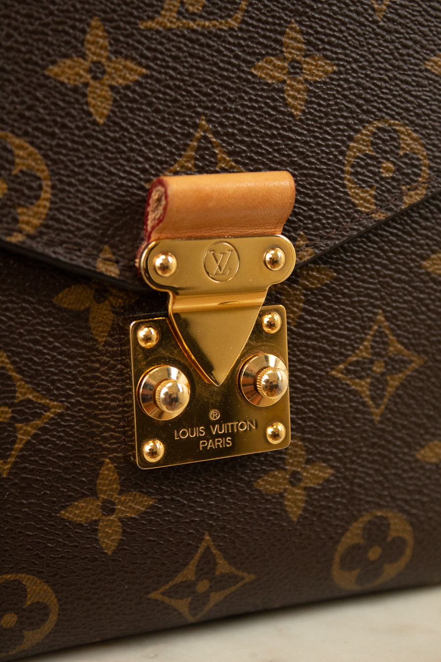 Close-up of a Louis Vuitton monogram handbag in brown coated canvas with gold-tone hardware and a tan leather strap tab, showing the Louis Vuitton Paris engraved clasp.