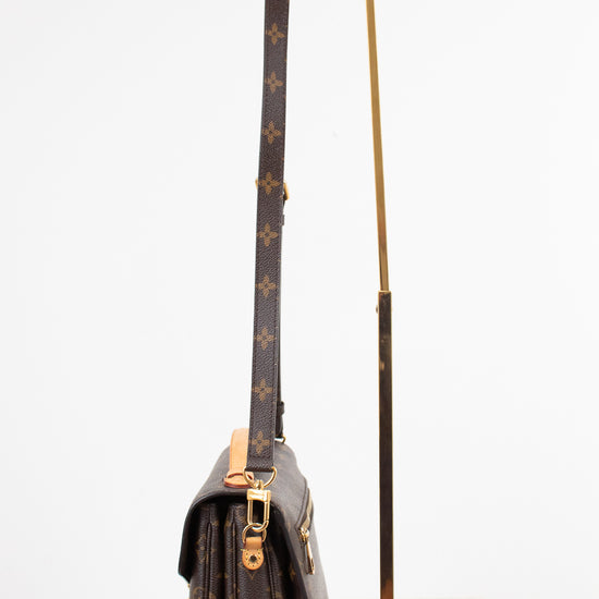 A Louis Vuitton monogram shoulder bag in dark brown coated canvas with gold-tone hardware, displayed next to a minimalist gold metal stand on a white marble surface against a white background.
