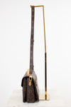 A Louis Vuitton monogram shoulder bag in dark brown coated canvas with gold-tone hardware, displayed next to a minimalist gold metal stand on a white marble surface against a white background.