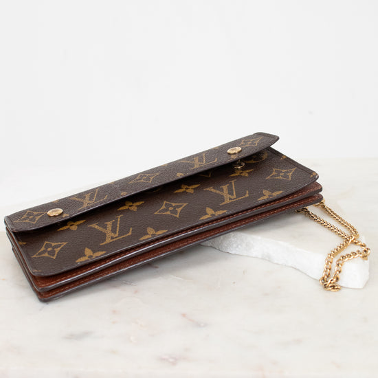 A Louis Vuitton monogram canvas long wallet or card holder with a gold-tone chain strap, resting on a white marble surface against a white background.