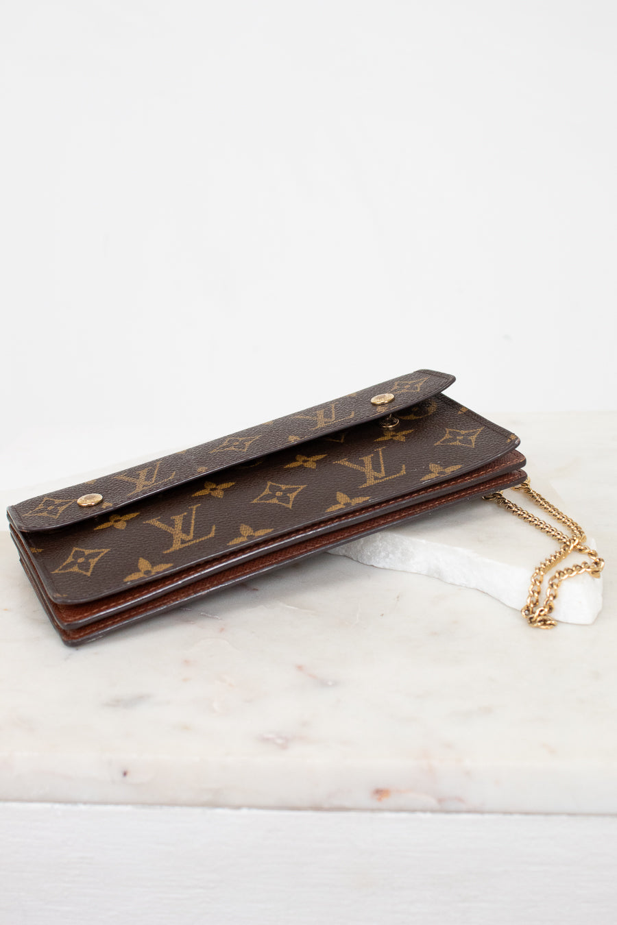 A Louis Vuitton monogram canvas long wallet or card holder with a gold-tone chain strap, resting on a white marble surface against a white background.