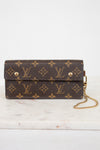 A Louis Vuitton monogram canvas clutch bag with a gold chain strap, resting on a white marble surface against a white background.