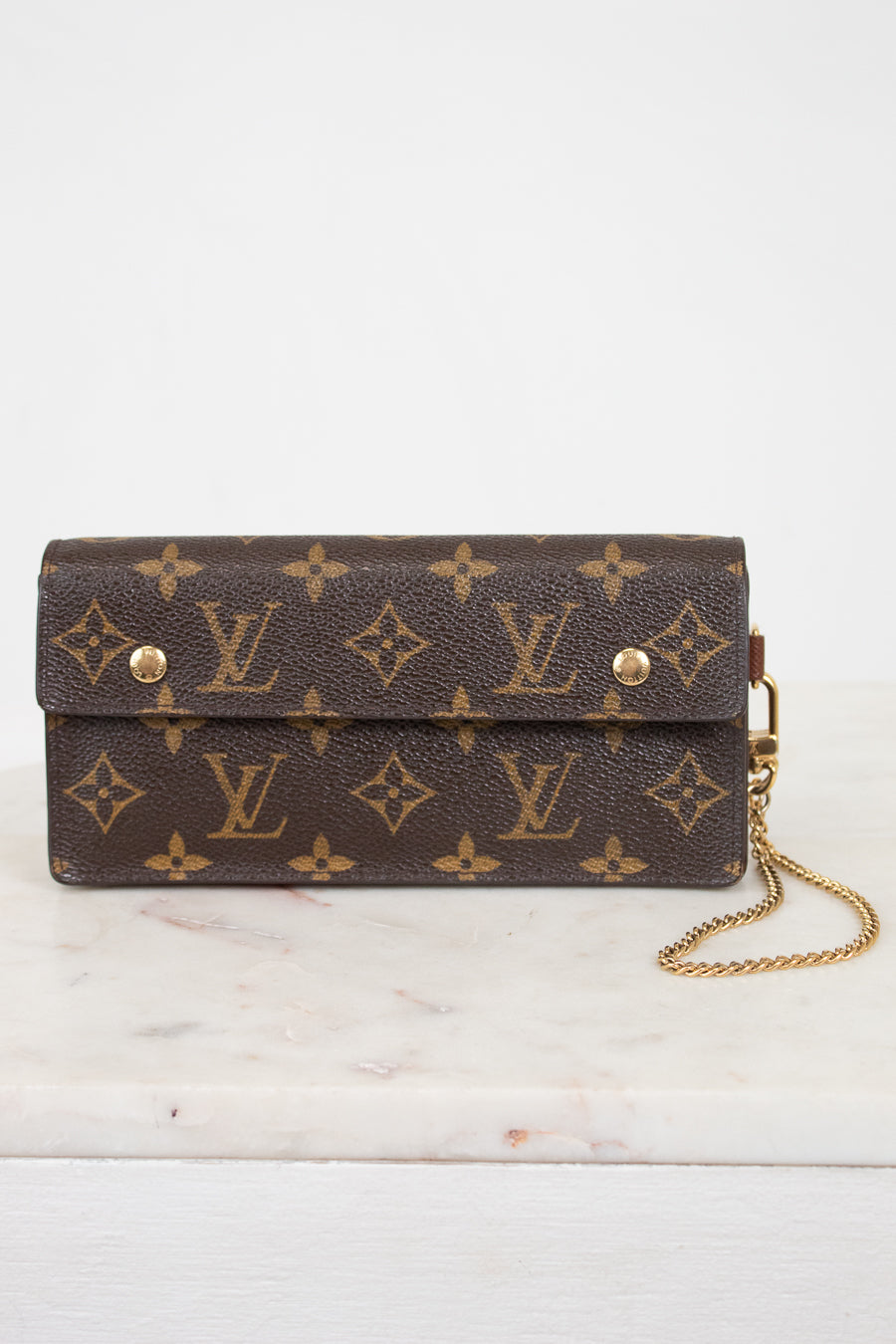 A Louis Vuitton monogram canvas clutch bag with a gold chain strap, resting on a white marble surface against a white background.